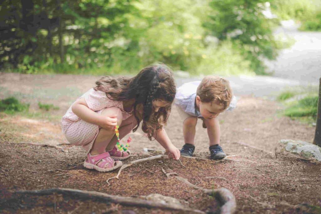 Preschool children exploring sticks and natural materials during outdoor summer holiday play.